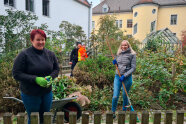 Studierende stehen mit Rechen, Gartenschere und Schubkarren im Hausgarten